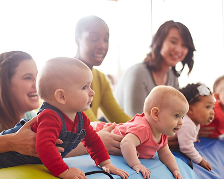 Bebés jugando y explorando sobre una superficie acolchada mientras adultos los acompañan en una actividad grupal de estimulación temprana.